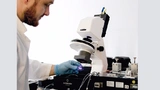 Ein Labormitarbeiter begutachtet den mikrofluidischen Chip. Rechts ist die Spritzenpumpe mit den Spritzen zu sehen. Um scharfe Bilder zu erhalten, arbeitet die Lichtquelle mit der Kamera synchron; jede Öffnung der Kamerablende löst einen Blitz aus. 