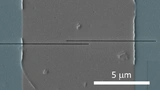 Rasterelektronenmikroskopische Aufnahme eines YBCO-Nanodrahts, geformt durch zwei horizontale Schnitte (schwarz) in einem YBCO-Film (grau). Der Maßstabsbalken (weiß) entspricht 5 Tausendstel Millimeter.
